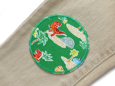Dinosaurier-Bügelflicken grün auf Kinderhose aufgebügelt, Baumwollstoff Ø 8 cm mit buntem Dino-Motiv Vulkane Palmen, Hosenflicken Kinderhosen reparieren Piratenbande Berlin